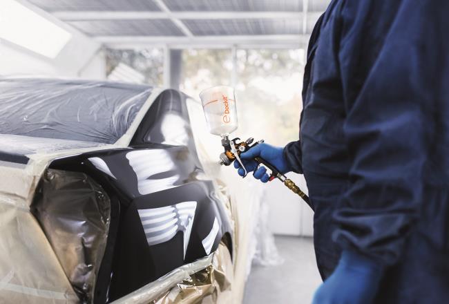 A man in overalls holds a Dockit capsule ready to apply clearcoat to a car exterior.