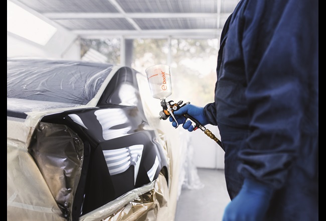 A man in overalls holds a Dockit capsule ready to apply clearcoat to a car exterior.